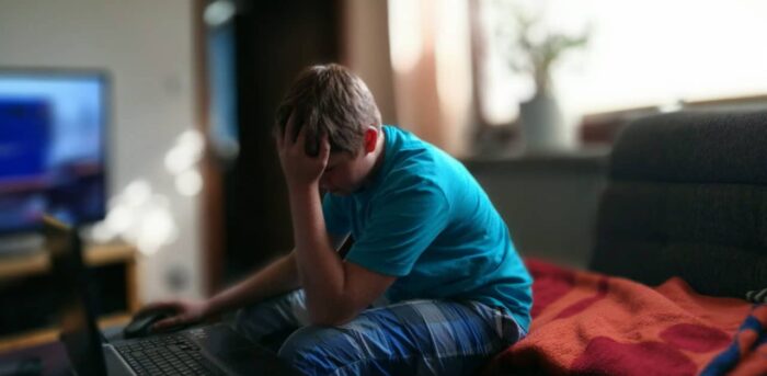 Un ragazzo con la testa tra le mani davanti al computer, in una stanza di casa, richiama il tema della legge sul bullismo e del cyberbullismo.