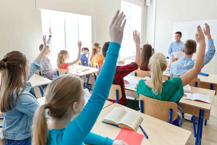 In una classe della scuola secondaria di secondo grado, studenti alzano la mano mentre il docente guida la lezione sul PEI e sull’ICF.